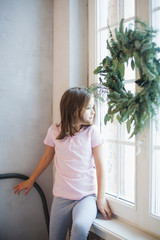 Girl standing near the window waiting for Santa Claus, Christmas wreath on the window, new year