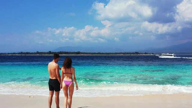 Young couple standing on white sand of exotic beach in front of blue turquoise sea where speed boats tracing on horizon on a cloudy morning in Bali