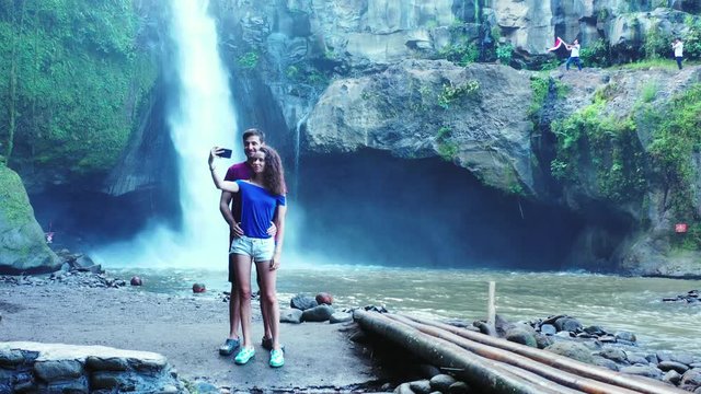 Happy couple taking picture with waterfall background on a beautiful natural park valley surrounded by big rocks in Thailand