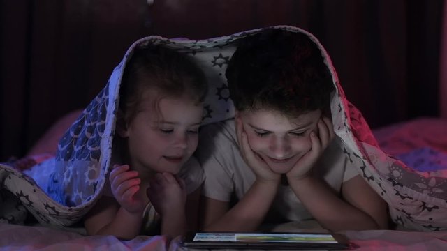 boy and a little girl covered with a blanket in the evening and watching cartoons or movies on a tablet in the bedroom on the bed. Brother and sister use a tablet to watch videos. Neon lighting.