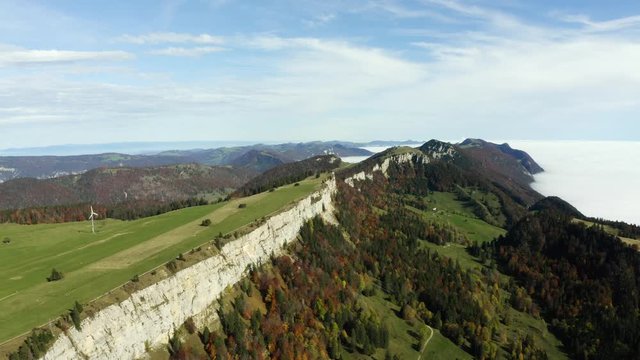 Epic aerial footage of autumn colored forest covered mountains at Grenchenberg in Switzerland with a sea of fog below in the valley, a long cliff visible and an electrical turbine on top
