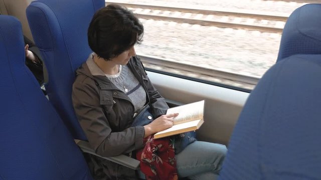 at the window of a moving train sits Caucasian middle aged woman brunette with short hair in a black jacket, blue jeans Reading a book runs his index finger over the lines side view from above