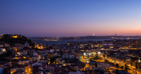 Scenic view of the downtown at dusk including Castelo de Sao Jorge, Alfama, Baixa and Bairro Alto districts, 25 de Abril Bridge over Tagus River and lit Cristo Rei monument in Lisbon, Portugal at dusk