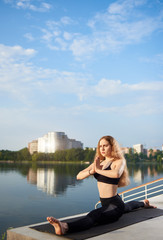 Fototapeta na wymiar Young woman practicing yoga on urban lake, wearing black top and pants. Relax in nature under blue sky. Sitting on the twine. Meditation. Residential background. Copy space