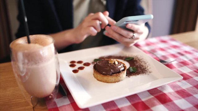 Young girl uses a smartphone, chooses photos of food, flips through web sitting in a cafe with dessert.