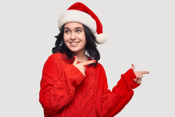 Pretty girl in a red sweater and Santa hat pointing at the copy space isolated over grey background