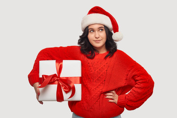 Brunette girl in a red sweater and Santa Claus hat holding white giftbox with red ribbon