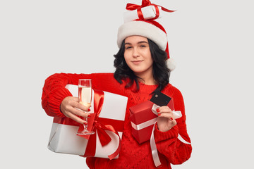 Brunette girl in a red sweater and Santa hat holding giftboxes, bank card and glass of champagne
