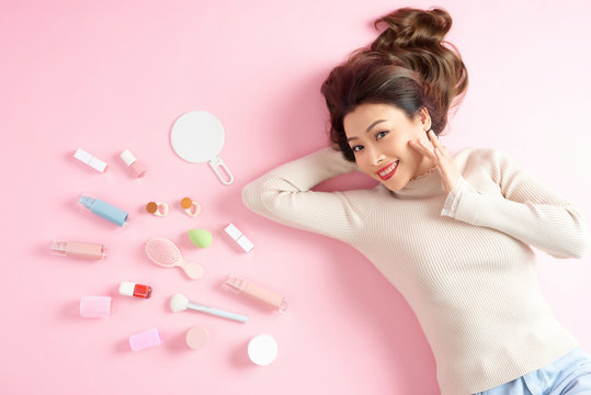Cheerful Asian woman lying on the pink floor with her cosmetic makup tools. Top view