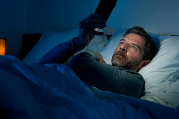 Fototapeta premium young attractive and relaxed man with blue eyes lying on bed late at night in dark and dim light networking on mobile phone or online dating in internet addiction