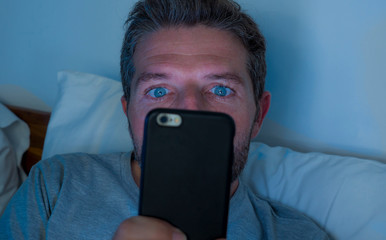 attractive and crazy internet addict man with blue eyes lying on bed late at night in dark and dim...
