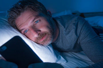Fototapeta na wymiar young attractive and relaxed man with blue eyes lying on bed late at night in dark and dim light networking on mobile phone or online dating in internet addiction
