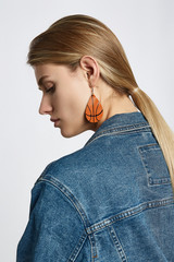 Profile portrait of a blond girl, wearing jean jacket. She has leather pop-art tearshaped basketball ball earrings. Girl is looking down.