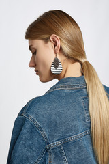 Profile portrait of a blond girl, wearing jean jacket. She has pop-art earrings with double leather tearshaped striped pendant. Girl is looking down.