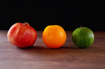 A red pomegranate, an orange orange and a small, green watermelon lie on a wooden surface. Horizontal arrangement, close-up, dark background.