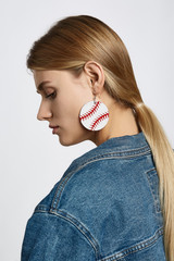 Profile portrait of a blond girl, wearing jean jacket. She has white leather pop-art baseball ball earrings. Girl is looking down.