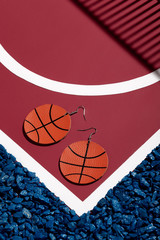 Object shot of white leather pop-art basketball ball earrings, lying on a red surface with white stripes and blue stones on borders. 
