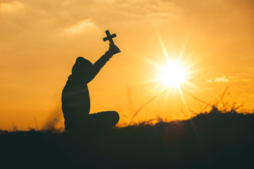 Man sit down and praying with raise the cross to sky at sunset background. christian silhouette concept.