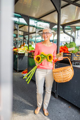 Senior Woman carying basket full of fresh vegetables and fruits on open market. View at senior woman with flowers at the market. Enjoying in sunny weekend morning. Caucasian ethnicity.