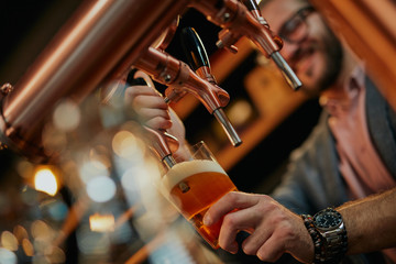 Close up of handsome caucasian bearded bartender standing in bar and pouring beer from tap. - obrazy, fototapety, plakaty