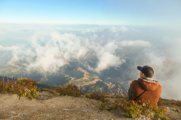 A young man sitting of the top of volcano Inerie in Bajawa, Flores, Indonesia. He is enjoying the beautiful view on volcanic island. There are some clouds below him. Discovering while travelling.