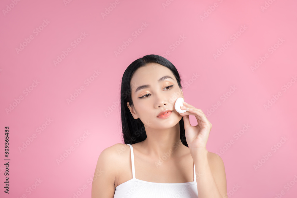 Wall mural Crop attractive young female cleansing face with a cotton pad on a pink background in studio