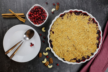 Crumble pie with cranberries and lingonberries in a white bowl, against a dark background decorated with a red napkin, cutlery, berries, nuts and cinnamon sticks, top view.