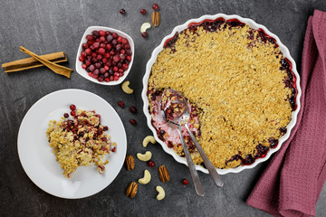 Crumble pie with cranberries and lingonberries in a white bowl, against a dark background decorated with a red napkin, cutlery, berries, nuts and cinnamon sticks, top view.