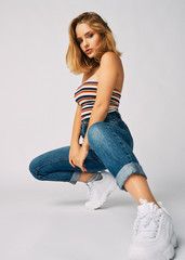 squatting young and athletic girl in jeans and a short T-shirt, beautiful and healthy skin, natural blond hair, empty background, delicate makeup,