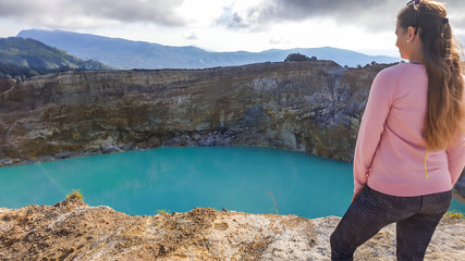 Naklejka premium A woman standing at the volcano rim and watching the Kelimutu volcanic crater lakes in Moni, Flores, Indonesia. Woman is relaxed and calm, enjoying the view on turquoise lake. Big overcast.