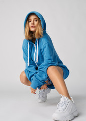 a young and athletic girl posing for photos in a large blue sweatshirt, beautiful and healthy skin, natural blond hair, empty background, delicate makeup,