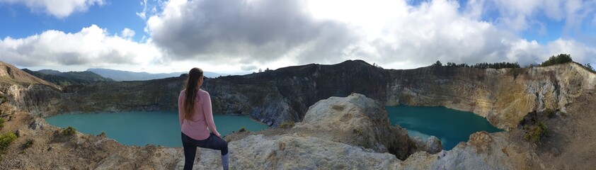 A woman standing at the volcano rim and watching the Kelimutu volcanic crater lakes in Moni, Flores, Indonesia. Woman is relaxed and calm, enjoying the view on turquoise lake. Big overcast..