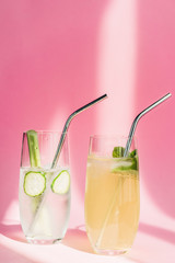 fresh lemonade in glasses with mint, ice, celery, straws and cucumber slices in sunlight on pink background