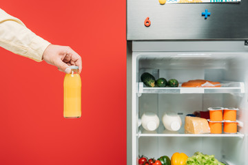 cropped view of man holding bottle of juice near open fridge with fresh food on shelves isolated on red