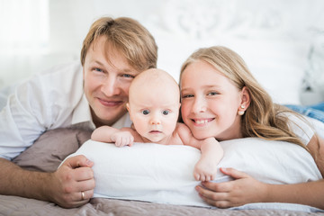 Obraz na płótnie Canvas Beautiful smiling young family mom dad elder daughter and newborn baby are lying on a large bed in a bright bedroom. Concept of friendly caucasian family