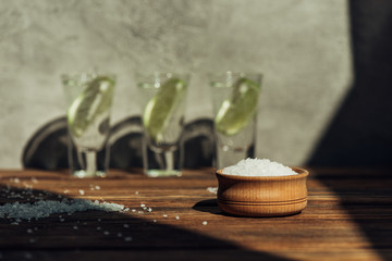 selective focus of salt in bowl and fresh tequila with lime on wooden surface in sunlight