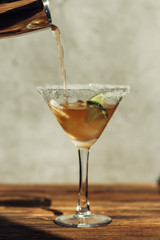 liquid pouring from shaker into martini glass with ice, lemon and lime decorated with sugar on wooden surface in sunlight