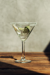 martini glass with ice, lemon and lime decorated with sugar on wooden surface in sunlight