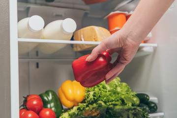 cropped view of woman taking bell pepper out from fridge with fresh food on shelves