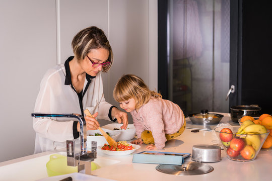 Mother preparing salad in the kitchen with a little girl sitting next to her who helps he