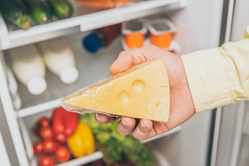 cropped view of man holding cheese near open fridge full of food
