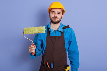 Closeup portrat it of painter wearing uniform,apron and protective helmet, holding roller isolated against blue studio background, bearded builder looking at camera, being ready for painting walls. - Powered by Adobe