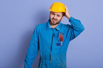 Studio shot of handsome young bearded Caucasian mechanic wearing blue work clothes and yellow hard hat, stands touching his head, having painful look, feeling exhausted after hard working day.
