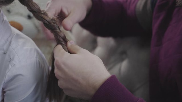Male Caucasian hands braiding pigtails. Father doing hairstyle for his brunette daughter. Fatherhood, care, lifestyle.