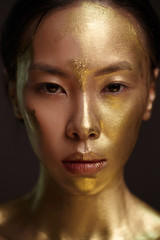 Beauty portrait of a beautiful аsian girl with gold paint on her face, against a dark background.