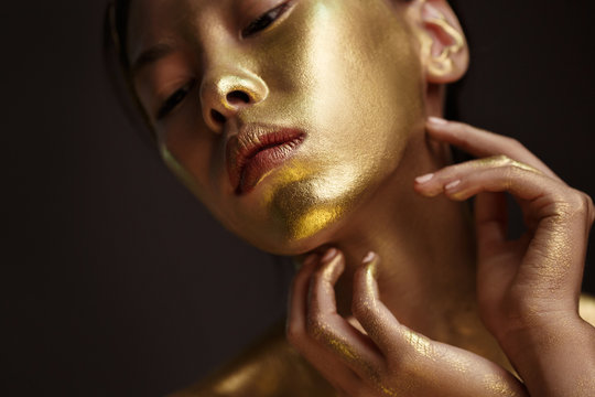Beauty portrait of a beautiful аsian girl with gold paint on her face, against a dark background.