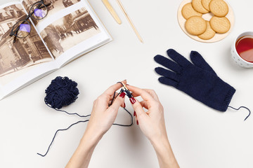Woman hands knit glove with bamboo spokes from blue wool yarn. Nearby knitted glove, book with vintage photos, glasses, cup of tea and cookies. Crochet work composition, close up. Needlework concept. 