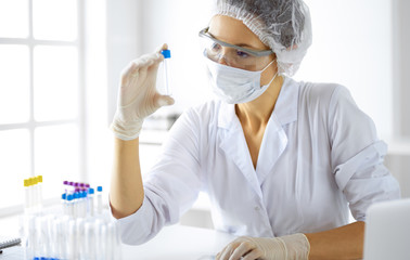 Professional female scientist in protective eyeglasses researching tube with reagents in...