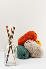 Vertical close up photo of multicolored wooden spokes in glass jar. On the background red, green, yellow and beige pastel colored balls of yarn. Selective focus. Needlework and knitting concept.