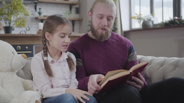 Close-up of happy Caucasian man reading book with teenage daughter. Cute girl with braided pigtails spending free time with father at home. Fatherhood, care, hobby, adolescence.